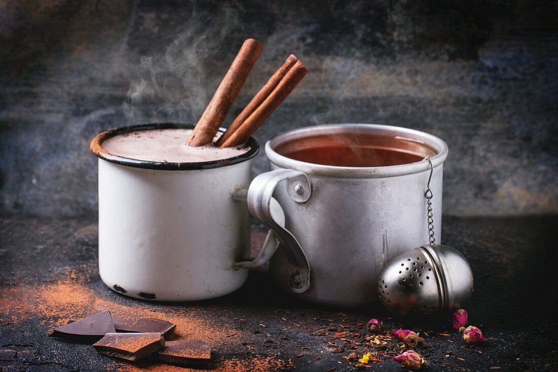 Heiße Schokolade mit Zimt und Kräutertee mit Tee-Ei und Rosenblüten in Emaillebechern