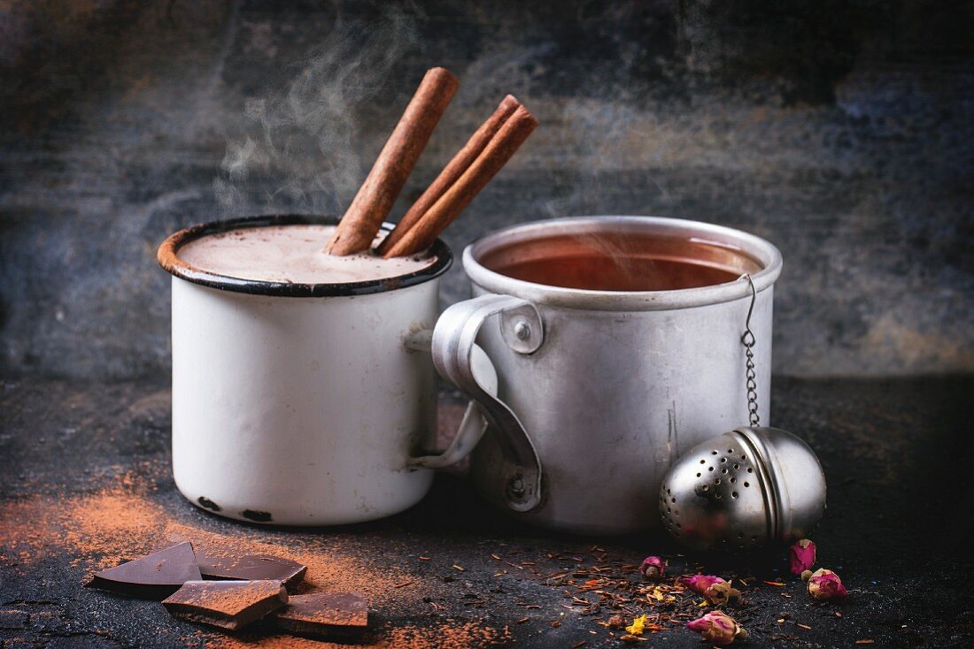 Two vintage mugs with tea and hot chocolate, served with dry tea, rose buds, chopped chocolate and cinnamon sticks