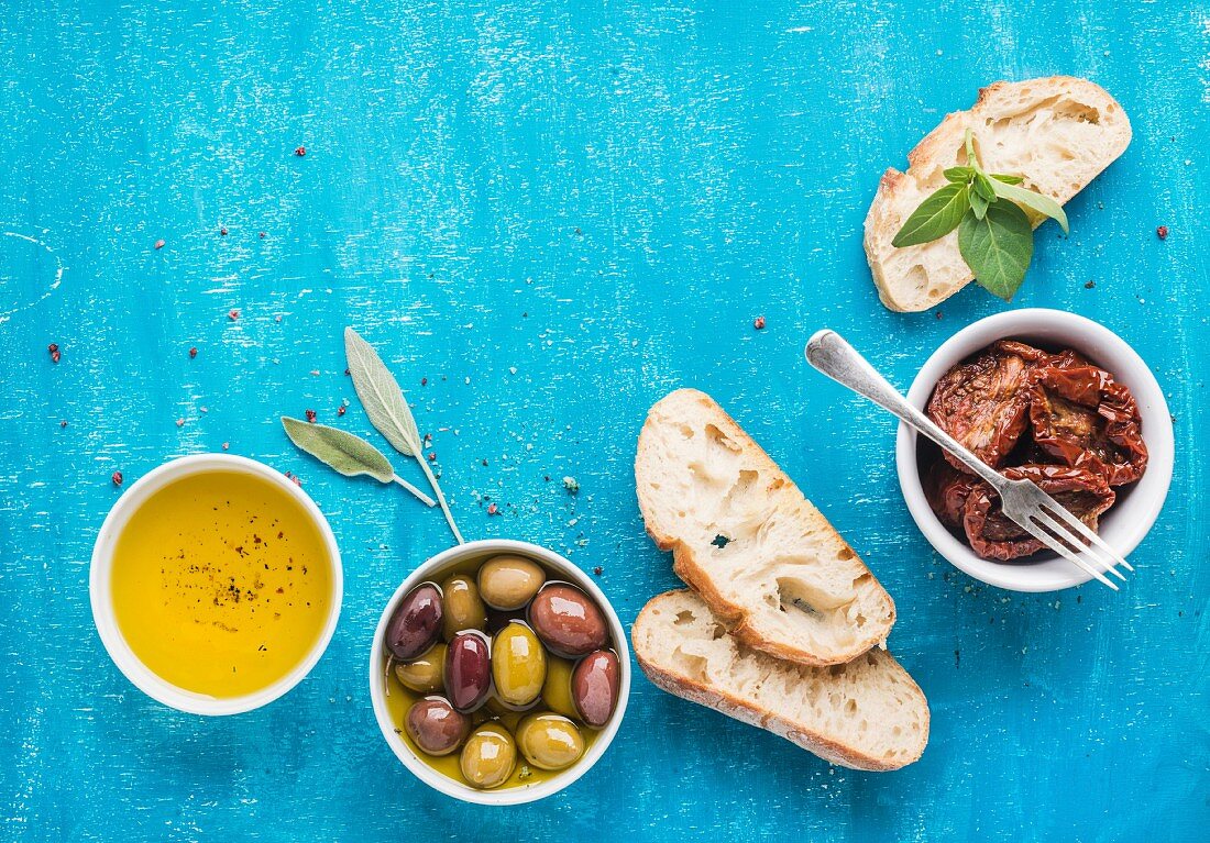 Pickled olives, oil, sun-dried tomatoes, herbs and sliced ciabatta bread over blue painted background