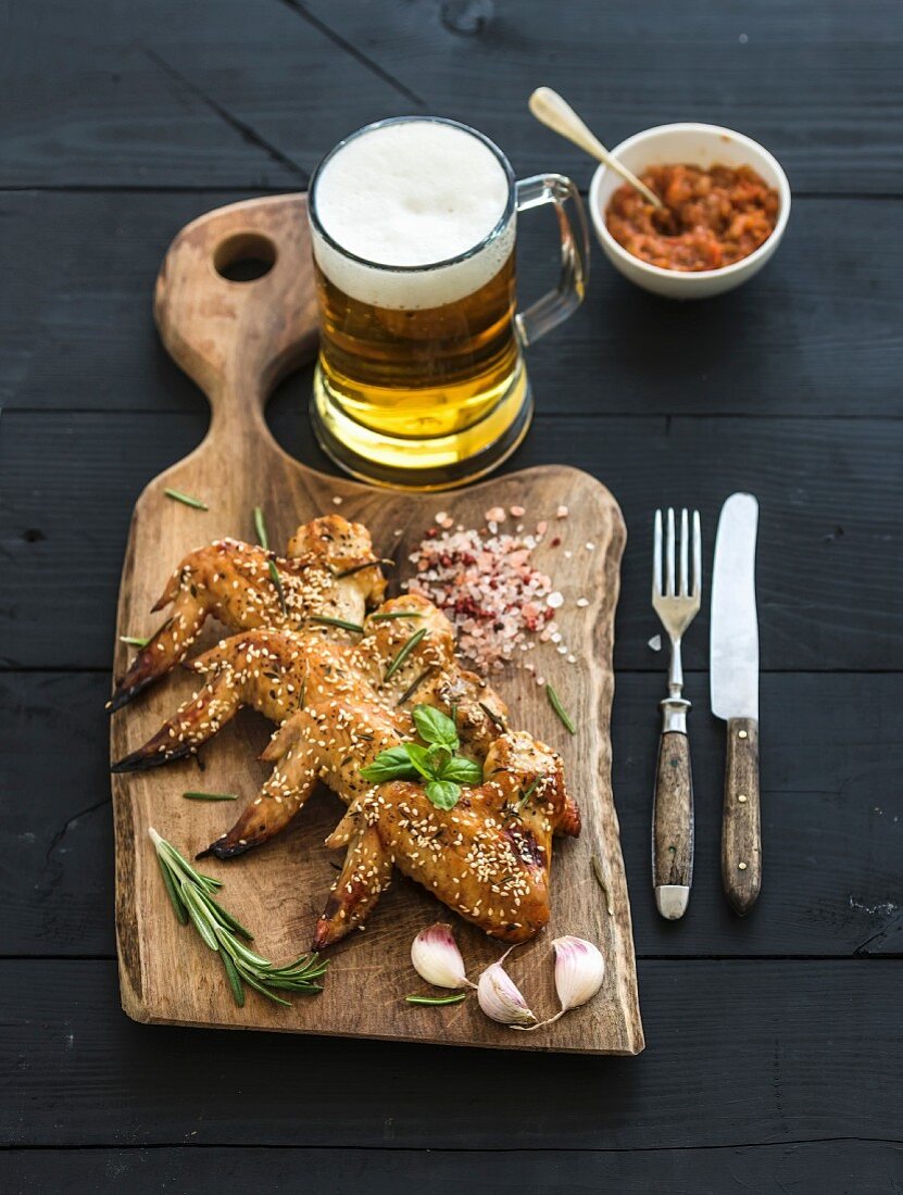 Gegrillte Hühnerflügel mit Sesam, Gewürzen und Kräutern, Glas Bier und Tomatendip auf rustikalen Holzbrett