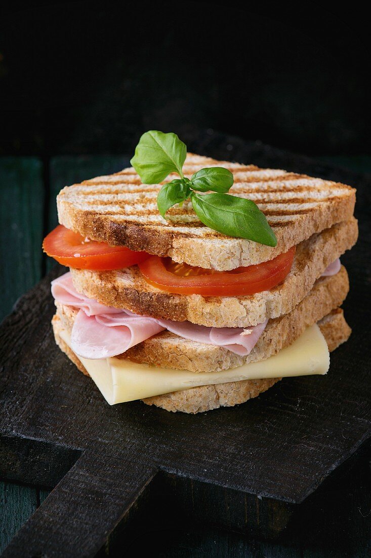 Whole grain grilled sandwich bread with ham, cheese and tomatoes on black wooden chopping board