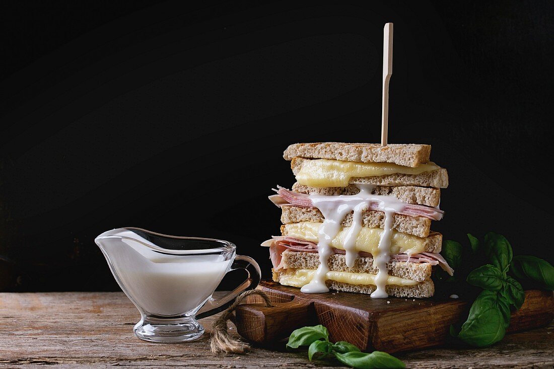 Whole grain grilled sandwich bread with melting hot cheese, ham, basil and pouring white sauce on wooden chopping board