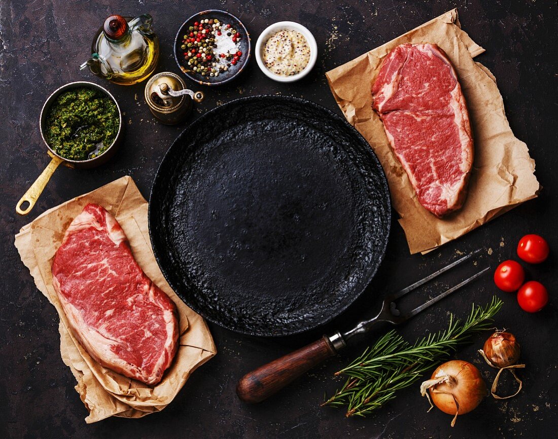 Raw fresh meat Striploin steaks with ingredients around frying pan on dark background