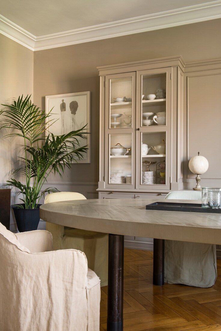 Loose-covered chairs around oval table and glass-fronted cabinet in dining room with pale grey walls