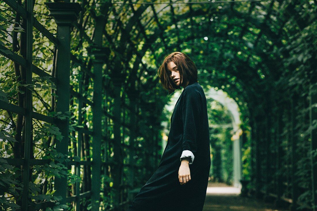A brunette woman wearing a black dress under a covered archway of foliage