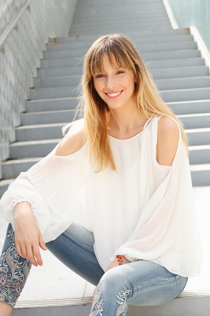 Blonde Frau in weißer Bluse und Jeans mit Blumenstickerei sitzt auf einer Treppe