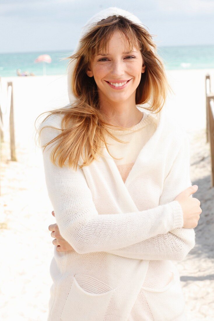 A blonde woman wearing a cream jumper and a white hooded cardigan on the beach