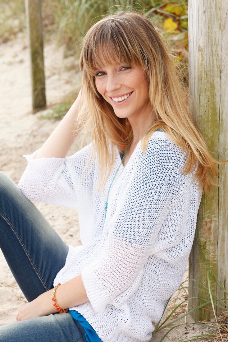 A blonde woman wearing a blue T-shirt, a white knitted jumper and jeans on the beach