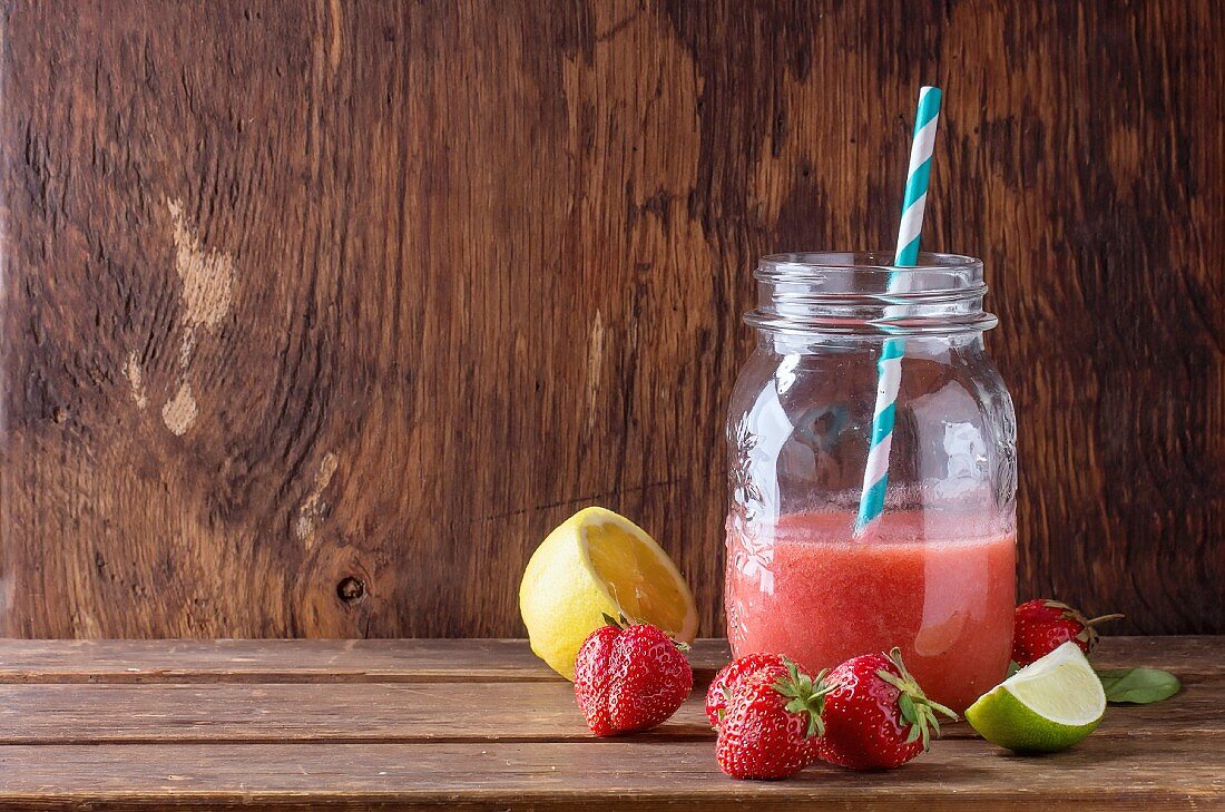Glass jar with red strawberry smoothie, served with fresh strawberries, lemon and lime over wooden table