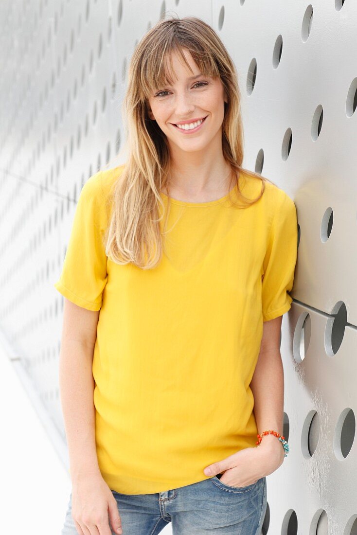 A blonde woman wearing a yellow T-shirt and jeans in front of a wall with holes