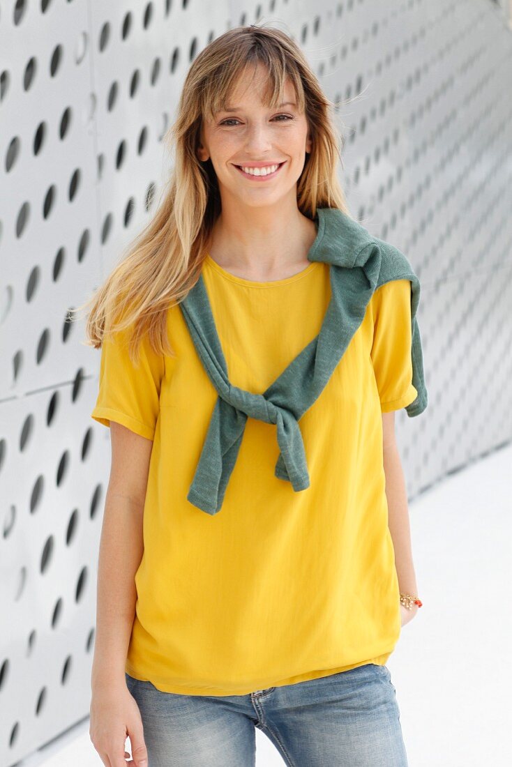 A blonde woman wearing a yellow T-shirt and jeans in front of a wall with holes