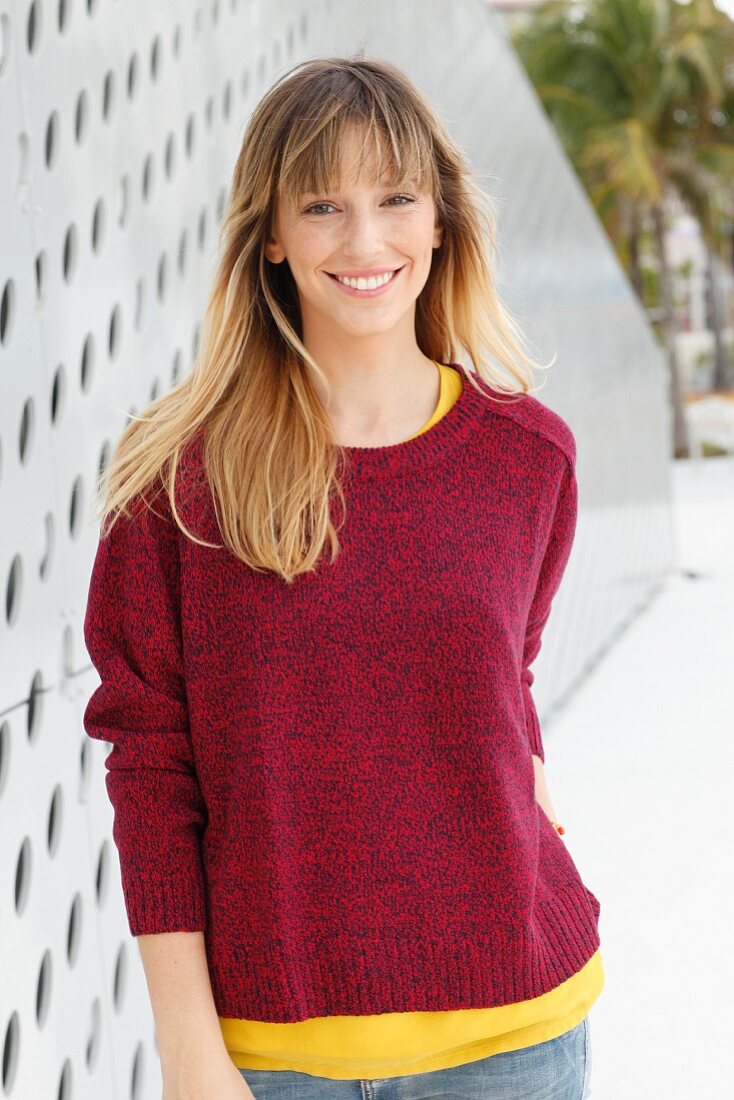 Blonde Frau in gelbem T-Shirt, rotem Strickpullover und Jeans vor Wand mit Löcher