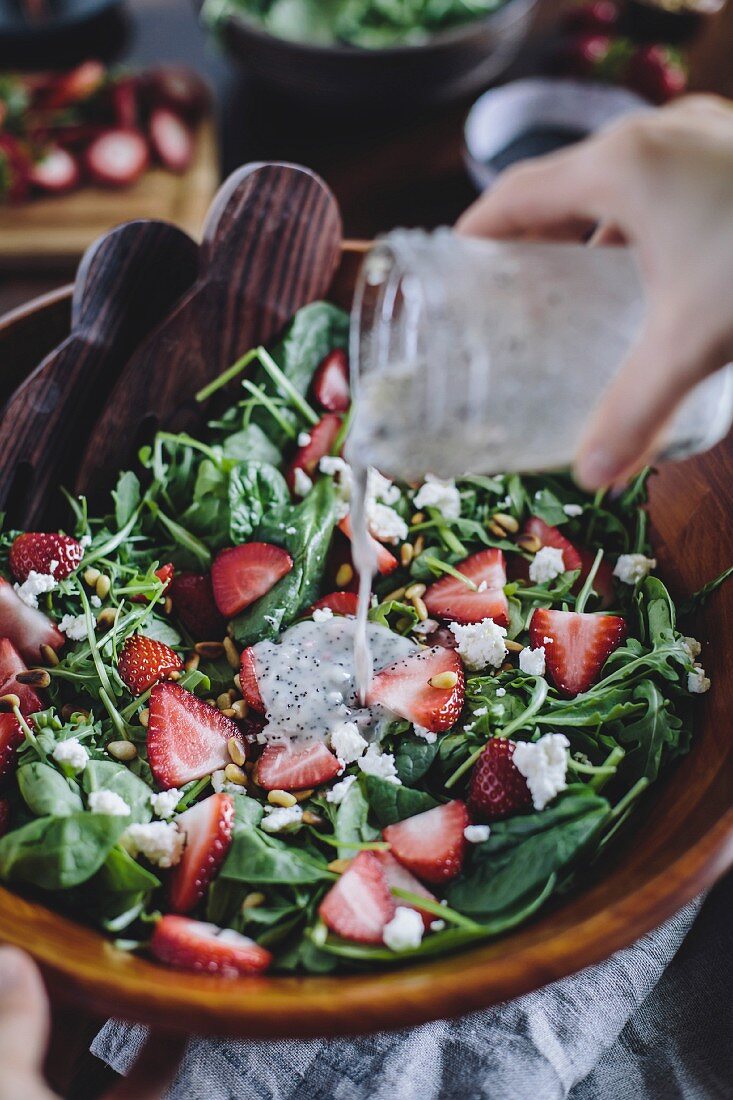 A woman is pouring poppy seed dressing into a strawberry and spinach salad