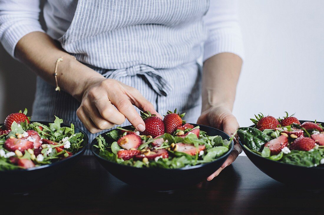 A woman is placing a strawberry into a bowl of strawberry spinach and arugula salad