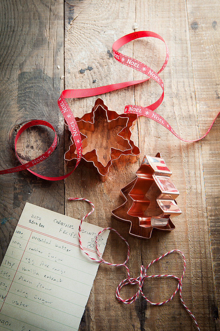 Copper christmas shaped cookie cutters on a rustic wooden surface with red Noel ribbon bakers twine and cookie recipe