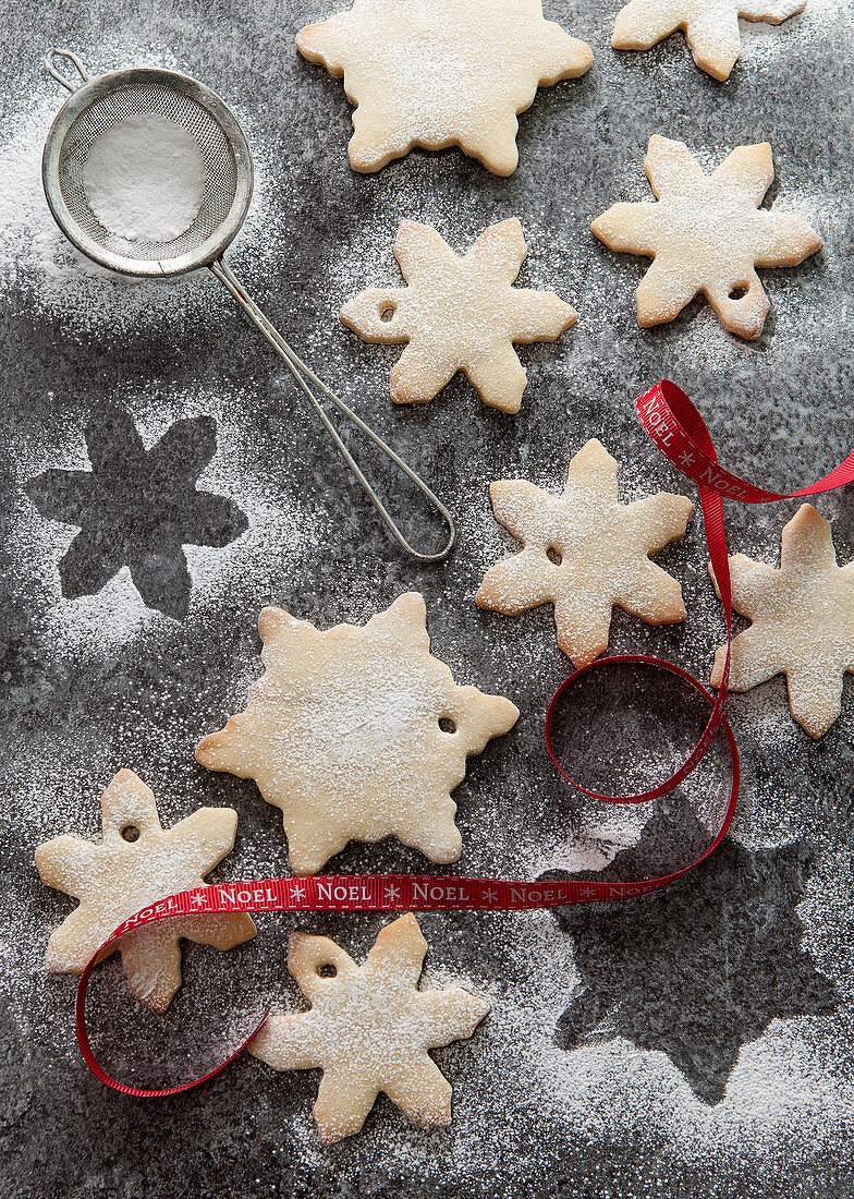 Two types of snowflake biscuits being dusted with icing sugar on a grey slate surface and an mini sifter and red Noel ribbon