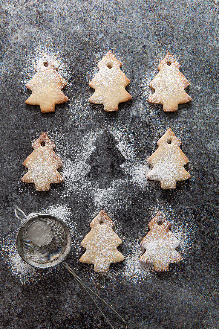 Rows of Christmas tree shaped biscuits dusted with icing sugar with one missing with just the outline remaining and a mini sifter filled with icing sugar all on a grey slate surface