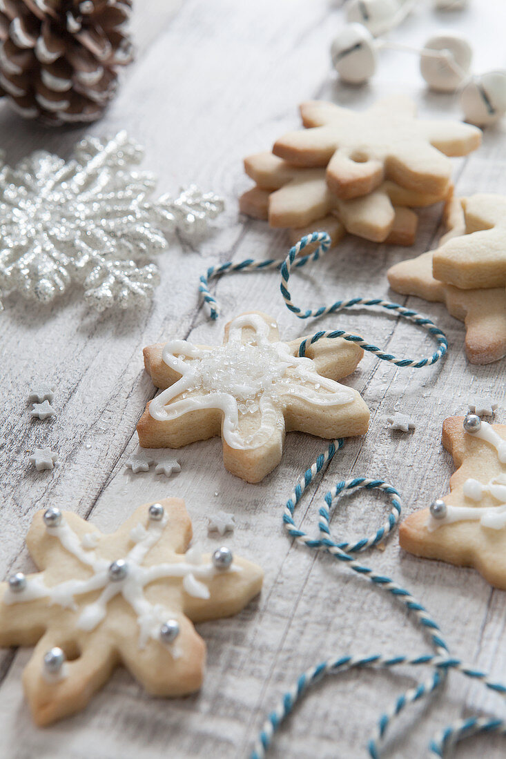 Iced homemade snowflake shaped christmas biscuits being treaded with blue and white bakers twine on a white wash wooden surface surrounded by Christmas tree decorations