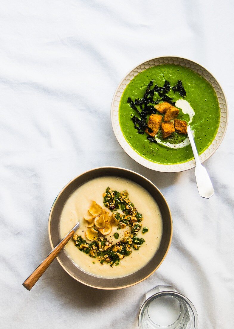 A bowl of parsnip soup with hazelnut-parsley gremolata and parsnip chips next to a bowl of parsnip and broccoli soup with kale and croutons