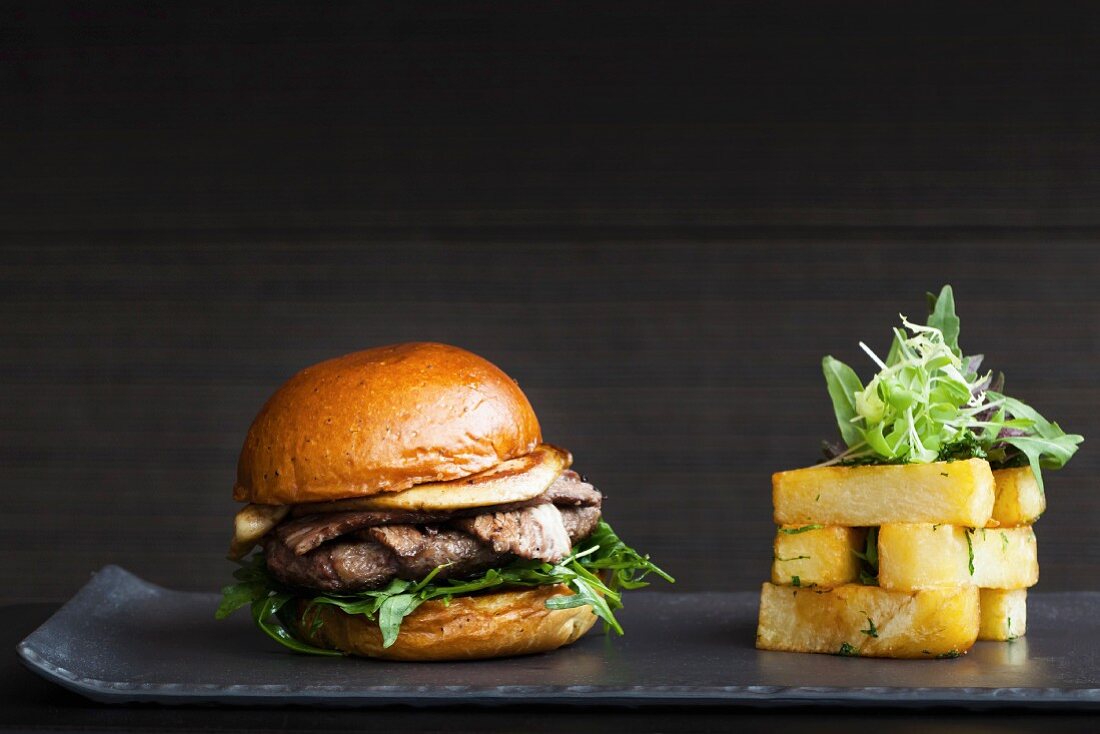 A juicy burger with bacon fois gras and lettuce in a bun served with a stack of chunky square cut chips on a black background