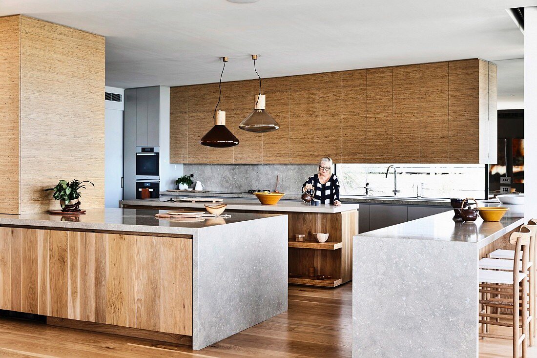 Kitchen units with wooden fronts and limestone worktop in open kitchen, woman in background