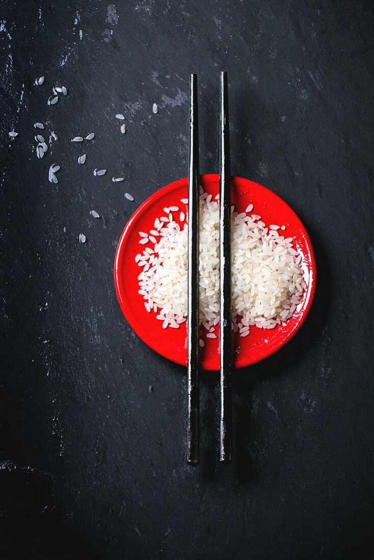 Uncooked rice on red plate with black wooden chopsticks over black surface