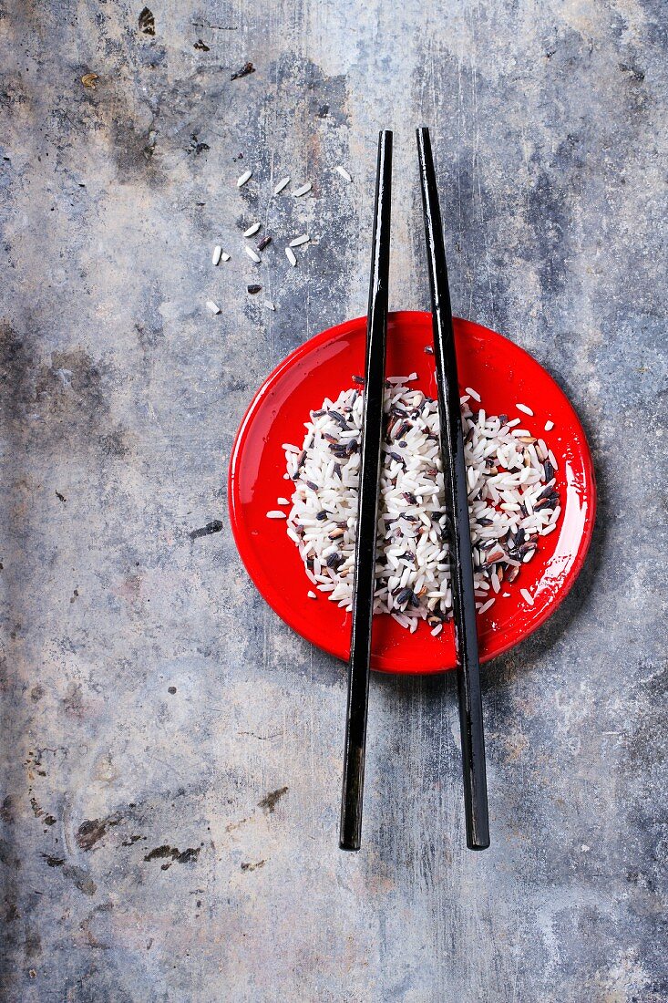 Ungekochter Reis mit Essstäbchen auf rotem Teller vor grauem Hintergrund