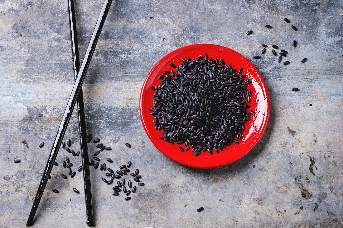 Uncooked black rice on red plate with black wooden chopsticks over gray tin surface