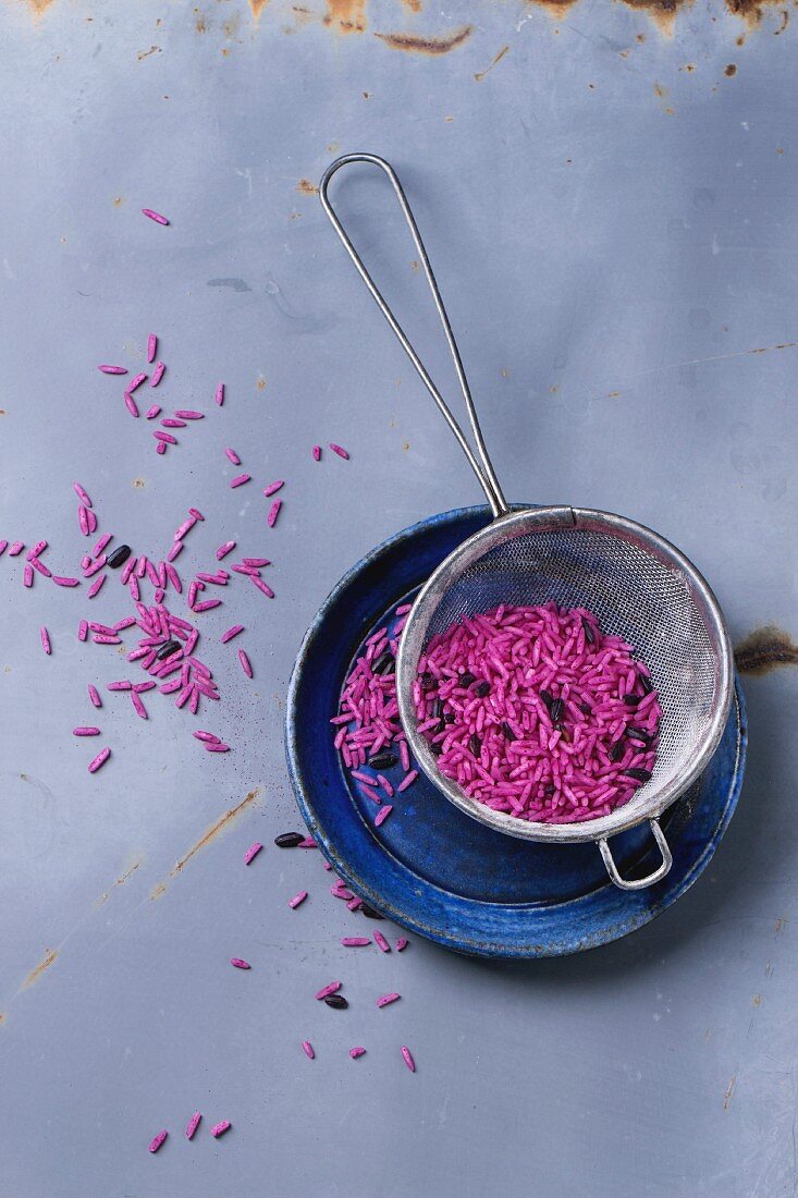 Uncooked pink and black rice on blue ceramic plate with sieve over gray metal surface
