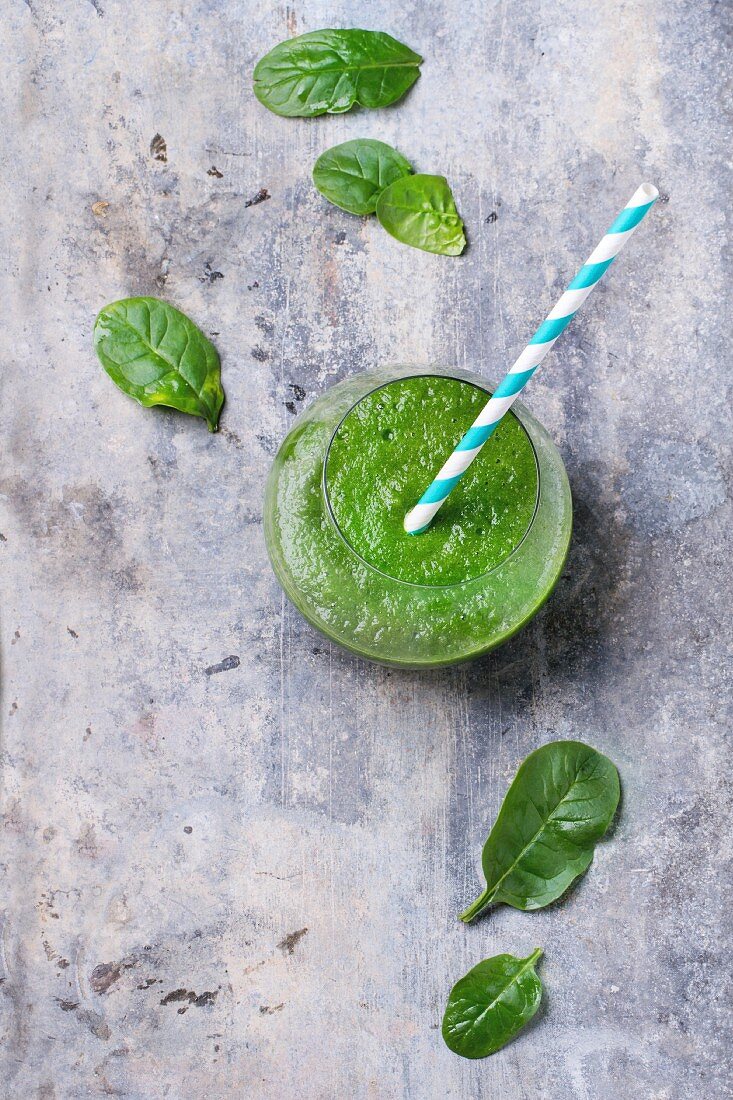 Glass of green smoothie, served with straw and baby spinach leaves over tin surface