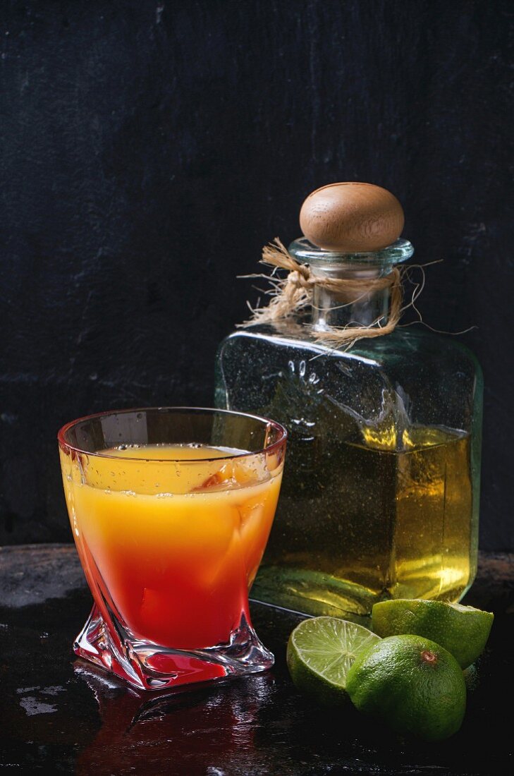 Glass of tequila sunrise cocktail, served with bottle of tequila anejo sliced limes over black background