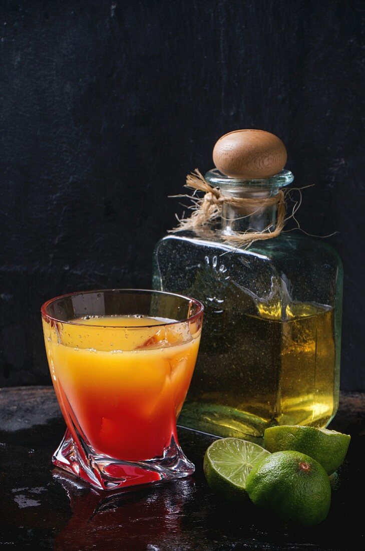 Tequila Sunrise im Glas, dahinter Tequila Anejo in Glasflasche