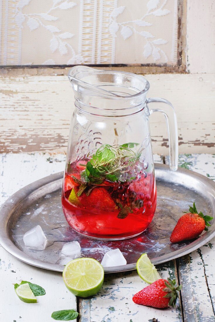 Glass jug of homemade strawberry lemonade, served with fresh strawberries, mint, lime and ice cubes