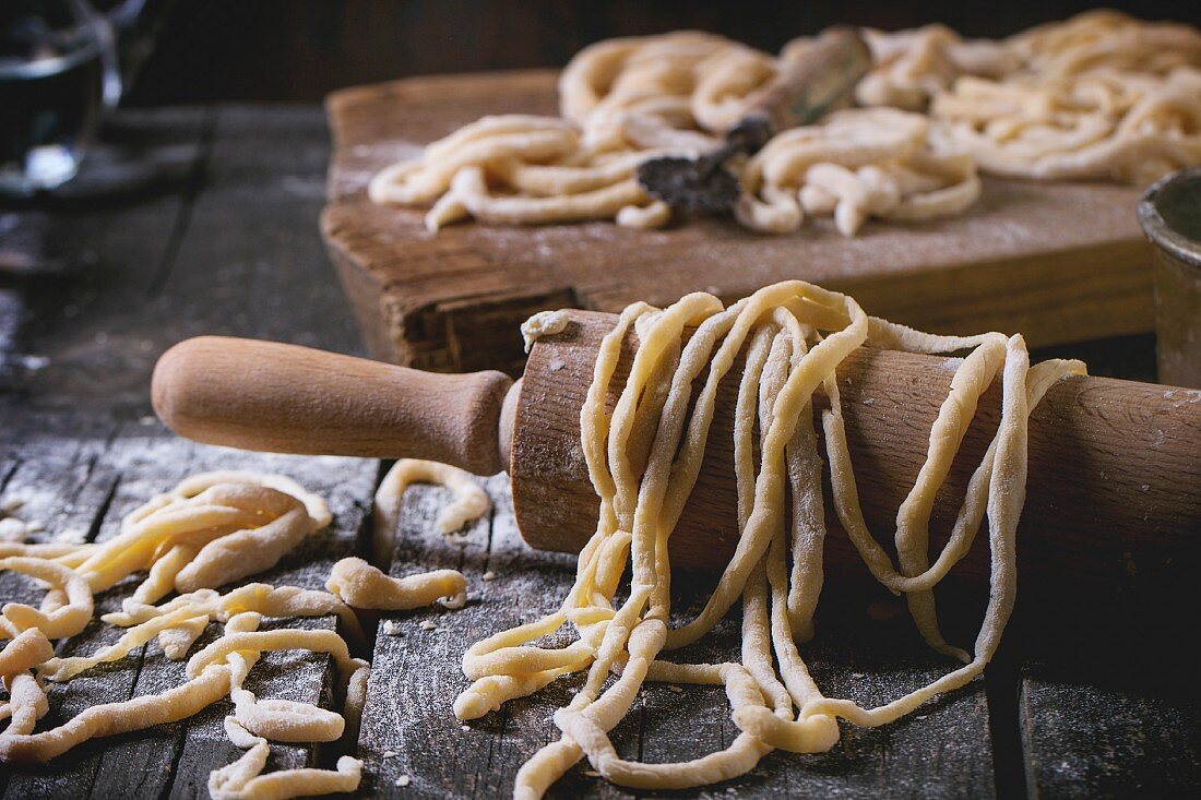 Fresh homemade pici pasta on wood chopping board over old wooden table with flour and rolling-pin