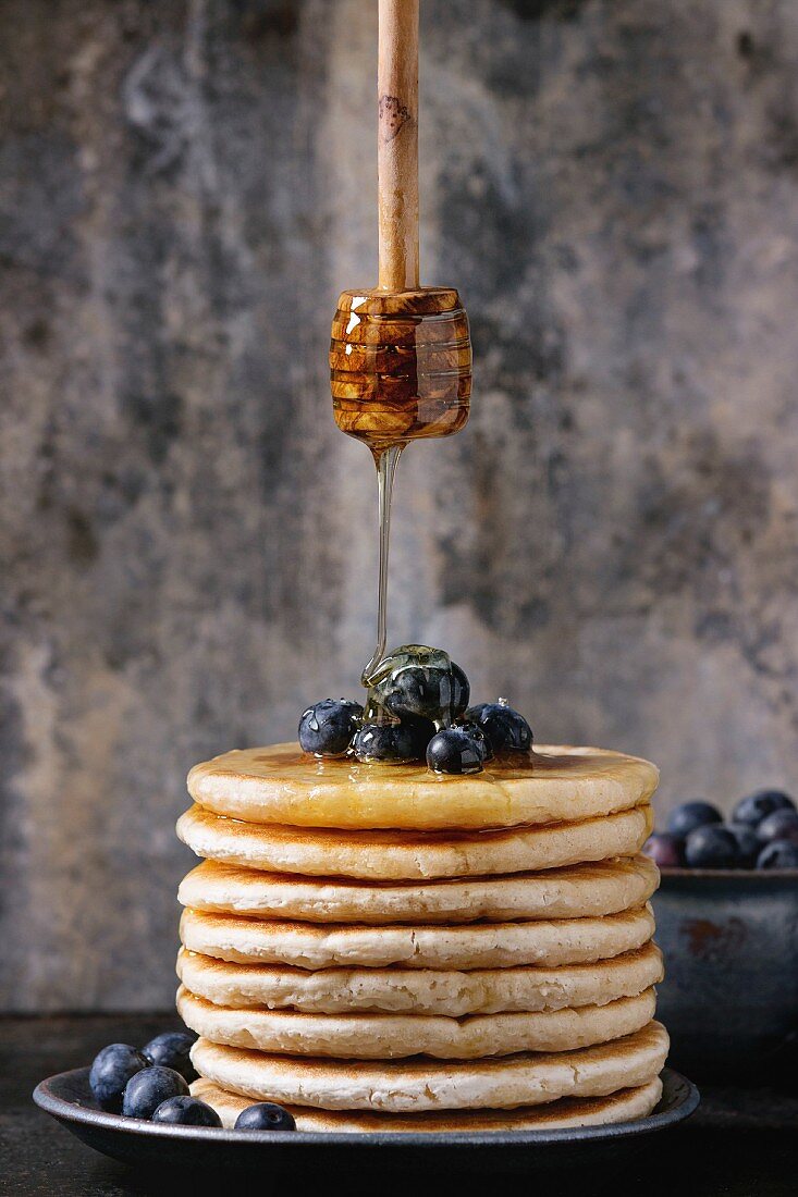 Pancakes with fresh blueberries in blue ceramic plate, and flowing honey from wooden honey dipper