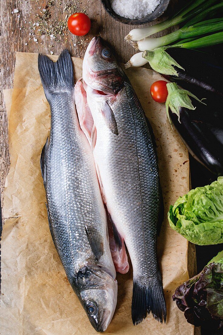 Two raw uncooked seebass fish on baking paper with sea salt, dry herbs and vegetables