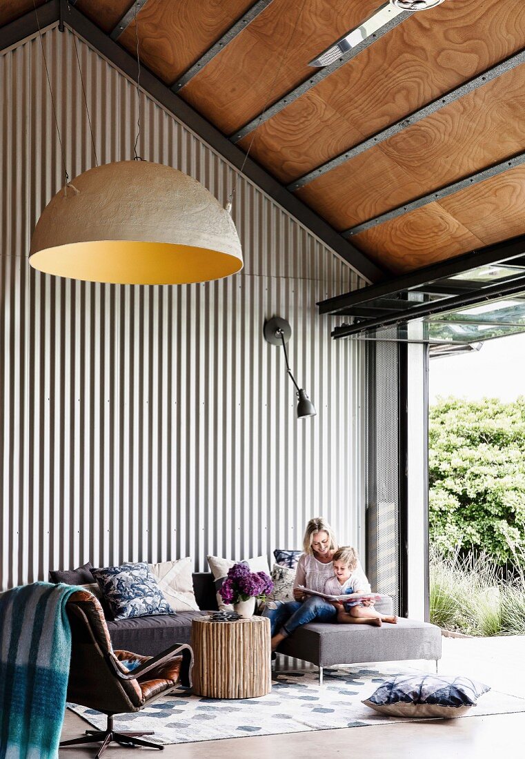 Woman and child on sofa in high living room with trapezoidal sheet cladding next to patio door in industrial style