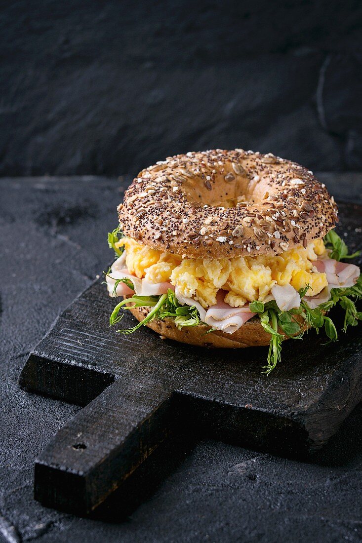 Whole Grain bagels with scrambled eggs, pea sprout and prosciutto ham on black wood chopping board