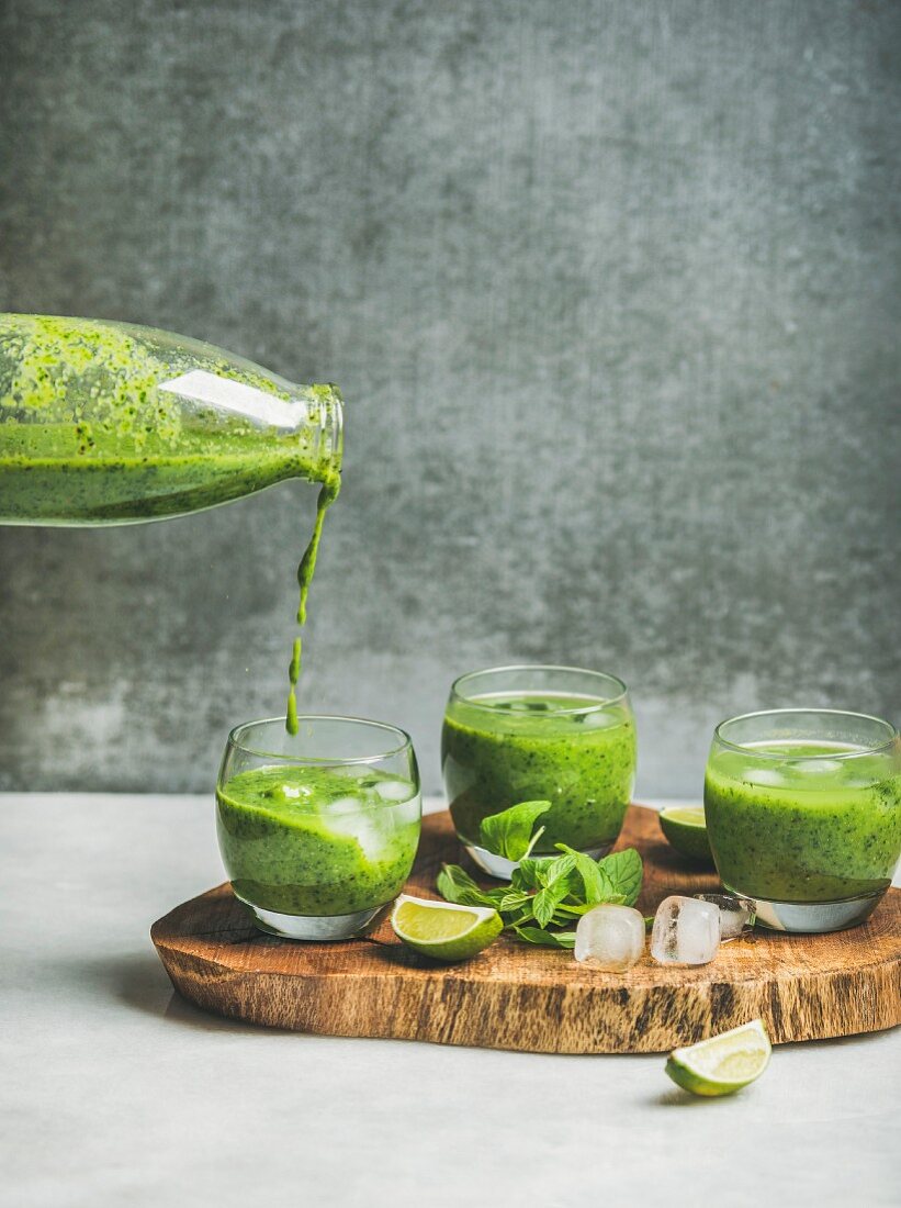 Fresh green smoothie in glasses and pouring from bottle with ice cubes, mint, lime on wooden board