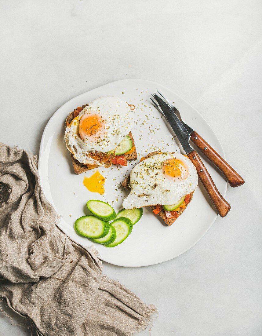 Breakfast toast with fried eggs with vegetables on white plate over grey marble background, top view