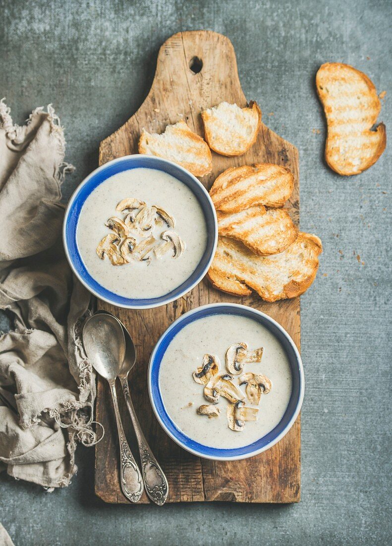 Creamy mushroom soup in bowls with toasted bread slices on rustic serving board, top view