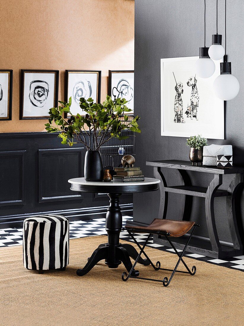 Round table, zebra-style pouf, leather folding chair, console table in front of black wall, black and white pictures in the background over black cassette cladding