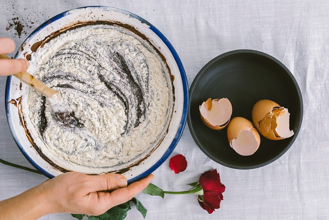 Hands stirring a mixture of flour, chocolate and cocoa powder in a mixing bowl with eggshells and a red rose