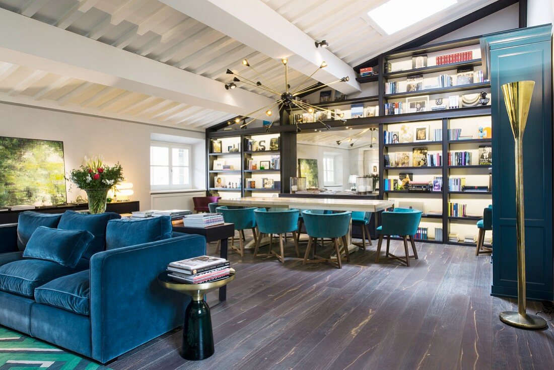 Blue sofa, long table, shell chairs and wall covered in shelving in open-plan interior
