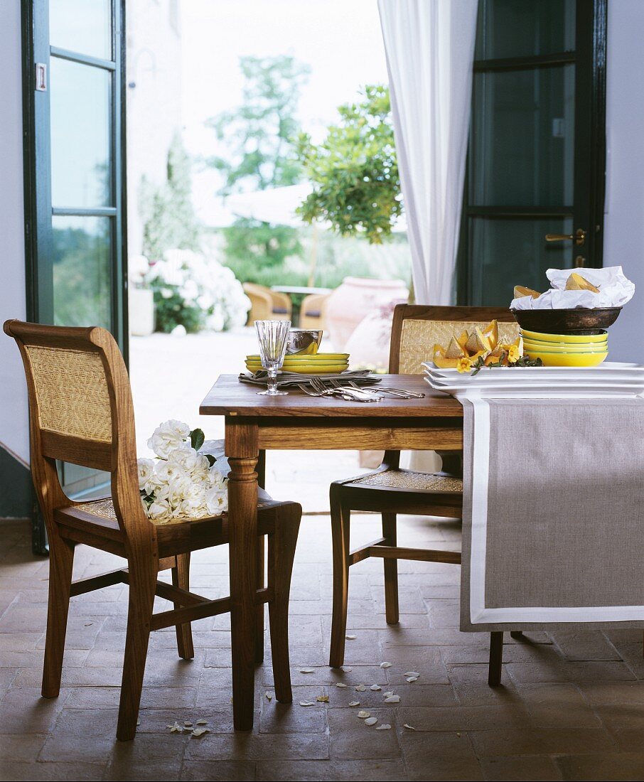 Crockery and cutlery on dining table and white roses on cane chair in front of open terrace doors