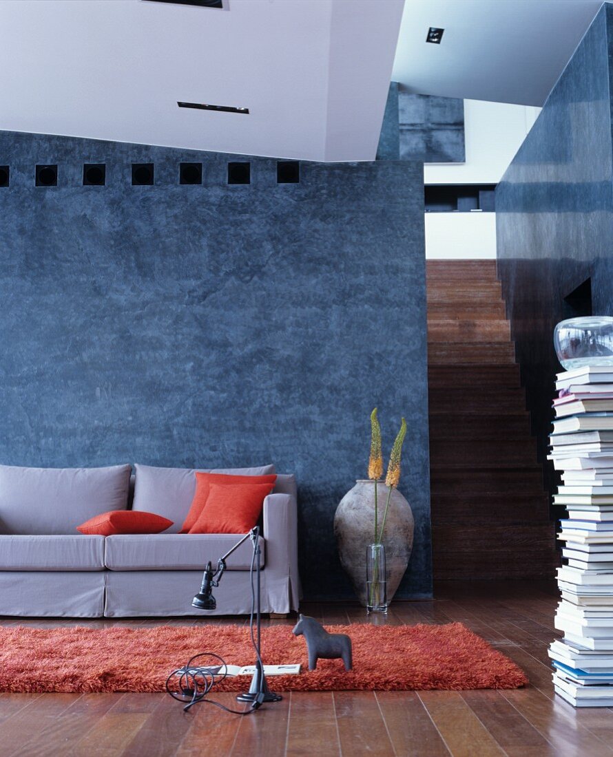Grey, loose-covered sofa with orange scatter cushions against grey-blue stucco lustro wall with stacked books and staircase to one side