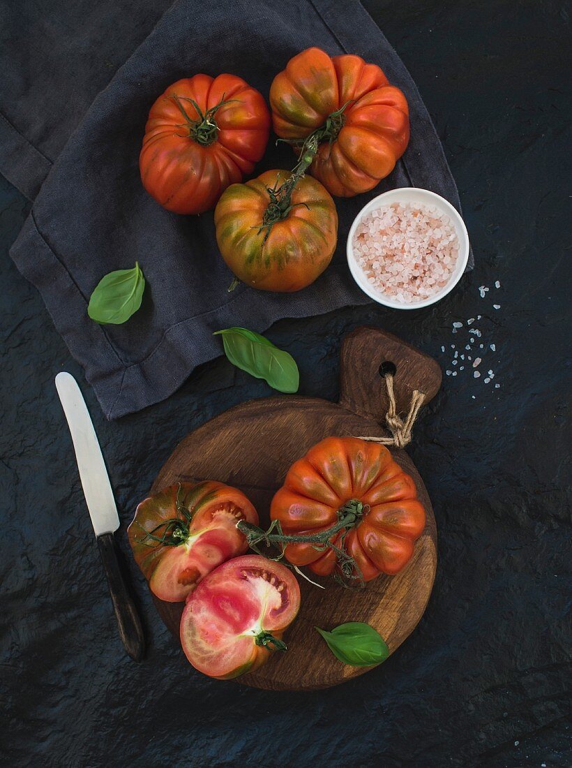 Fresh ripe hairloom tomatoes and basil leaves on rustic wooden board over black stone background