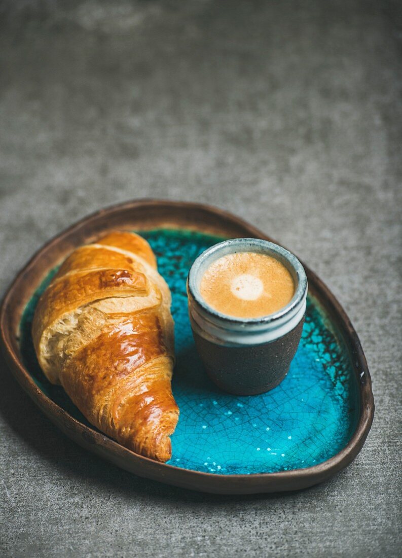 Cup of espresso coffee and croissant in turquoise blue ceramic tray over grey concrete background