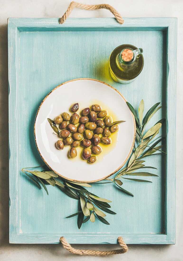 Eingelegte grüne Oliven mit Olivenölflasche und Olivenzweigen auf Holztablett (Aufsicht)