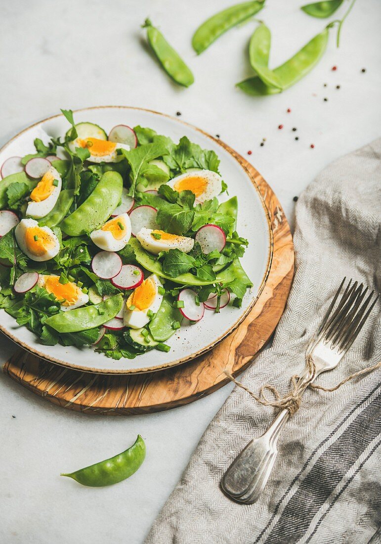 Healthy spring green salad with radish, boiled egg, arugula and  green pea in white plate on olive tree wood board